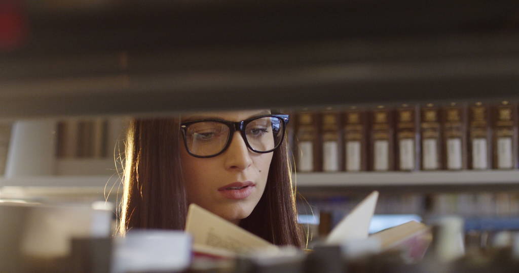 Młoda atrakcyjna kobieta studentka w okularach przewracać strony w książce wzięła się od książek na półkach w bibliotece. Portret. Z bliska. Wewnątrz - Zdjęcie, obraz