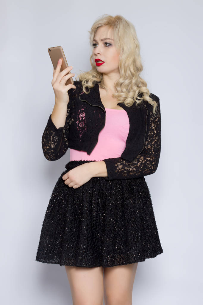 Porträt einer schönen jungen blonden Frau mit lockigem Haar in einem schwarzen Spitzenanzug mit einem Handy in der Hand - Foto, Bild