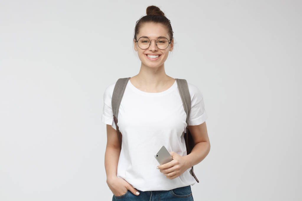 Jovem mulher de tshirt branca e óculos transparentes isolados em fundo cinza, sorrindo, de pé em mãos em bolsos pose
 - Foto, Imagem