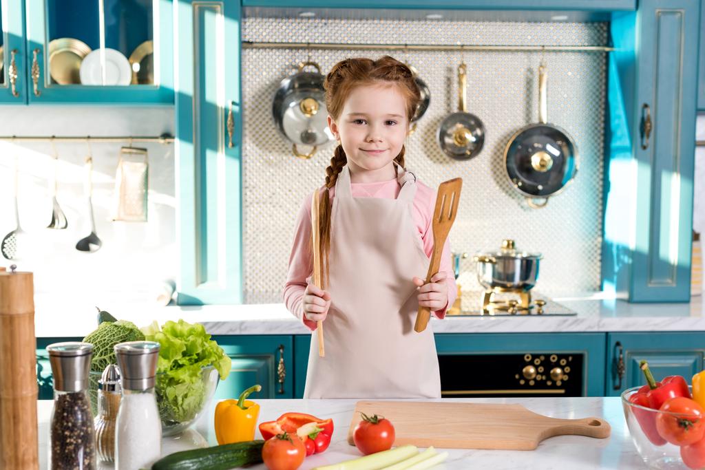 criança adorável em avental segurando utensílios de madeira e sorrindo para a câmera na cozinha
 - Foto, Imagem