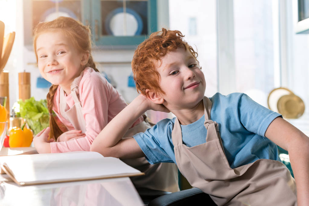 adorables niños en delantales sonriendo a la cámara mientras cocinan con libro de cocina en la cocina
 - Foto, imagen