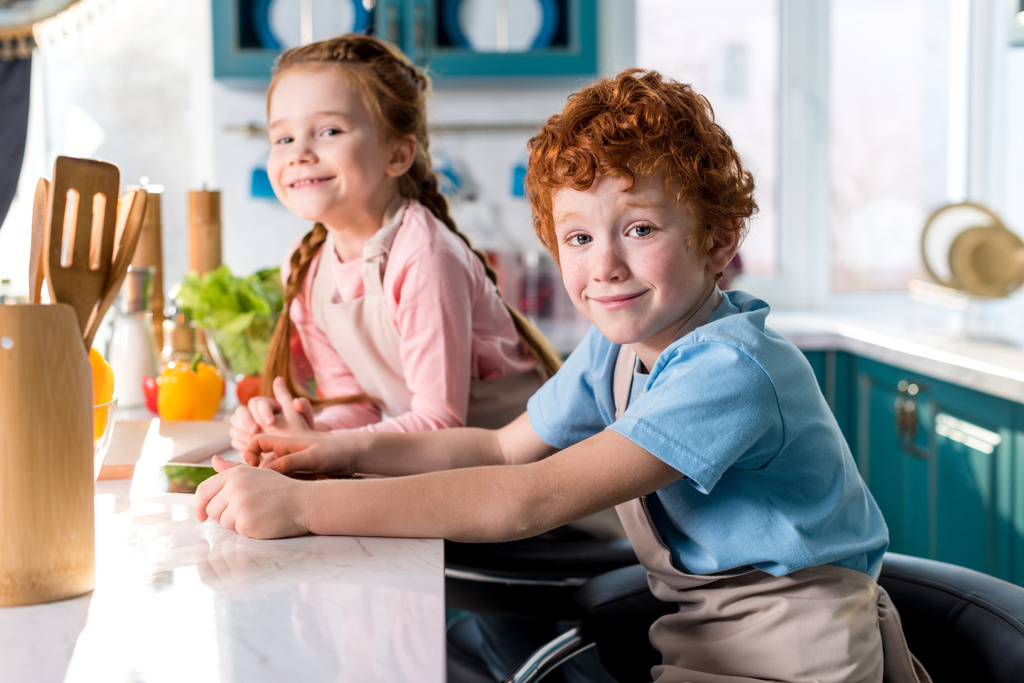 очаровательные счастливые дети улыбаются в камеру во время приготовления пищи вместе на кухне
 - Фото, изображение