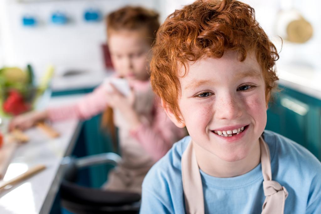 χαριτωμένο μικρό αγόρι χαμογελά στη φωτογραφική μηχανή, ενώ ο φίλος πίσω από το μαγείρεμα στην κουζίνα - Φωτογραφία, εικόνα
