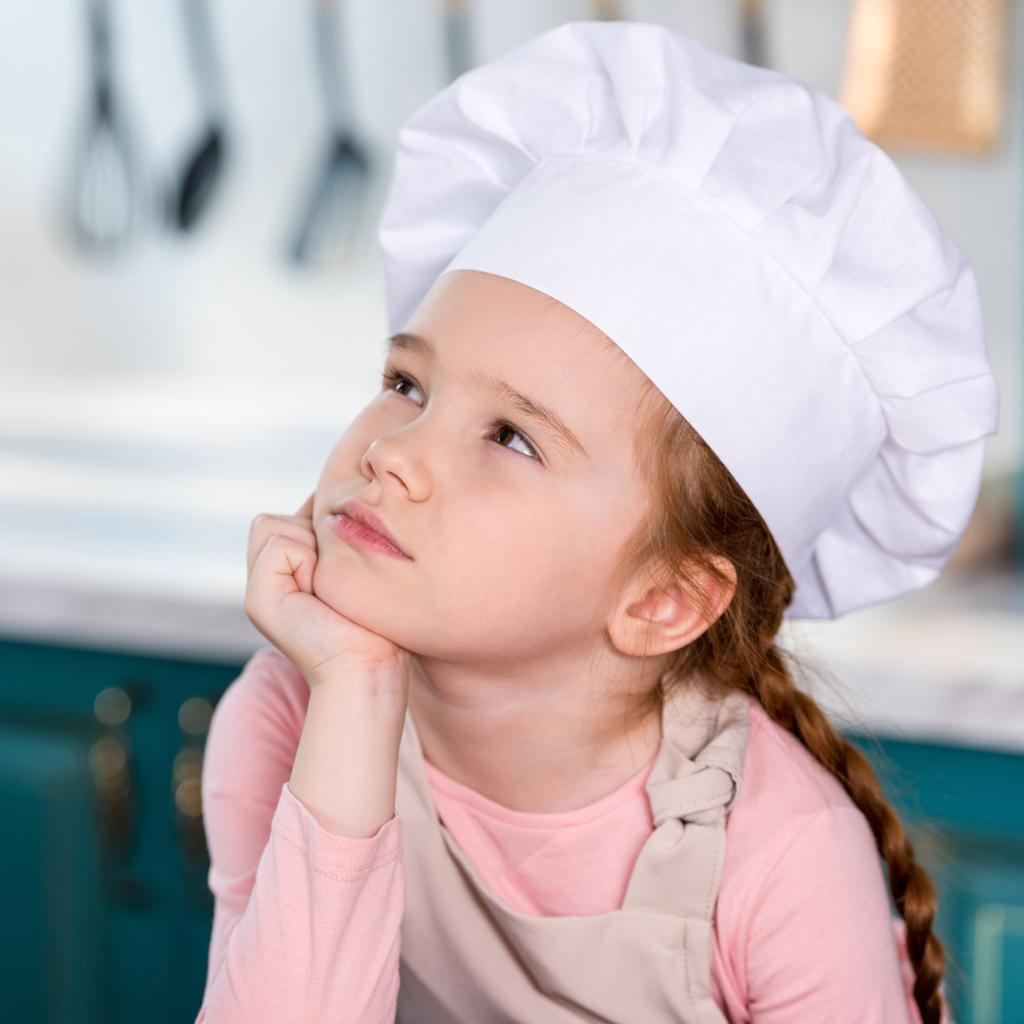 стомлена маленька дитина в капелюсі шеф-кухаря тримає руку на підборідді і дивиться на кухню
 - Фото, зображення