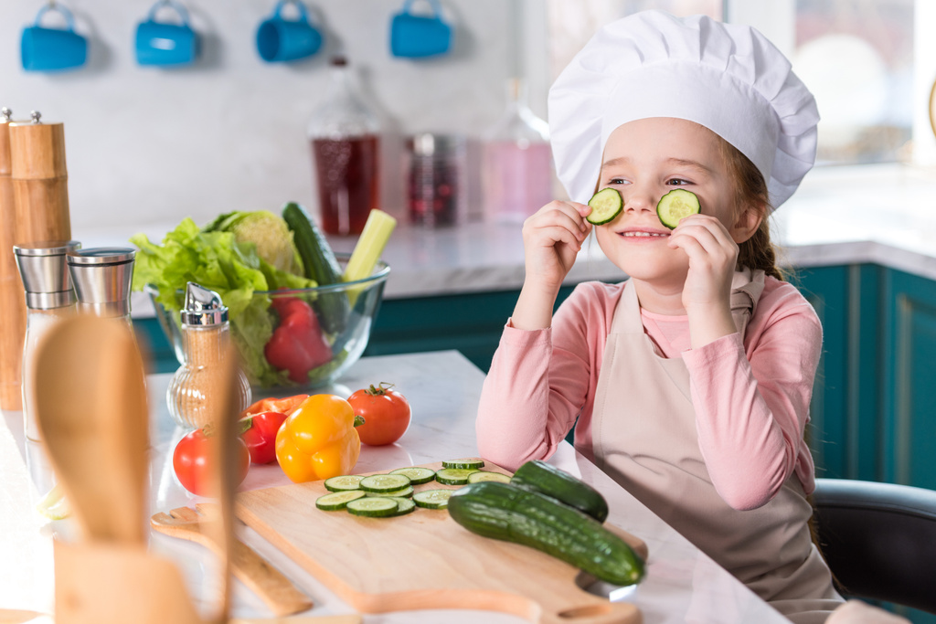 ребенок в шляпе шеф-повара и фартуке держа куски огурца во время приготовления пищи на кухне
 - Фото, изображение