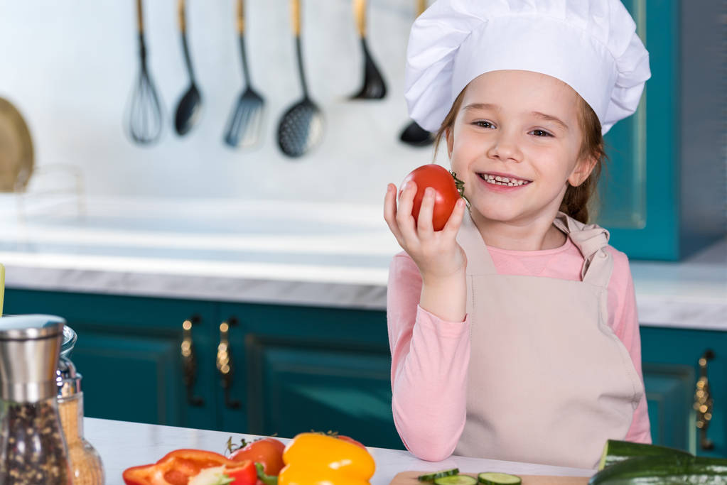 adorable niño en sombrero de chef sosteniendo tomate y sonriendo a la cámara en la cocina
 - Foto, imagen