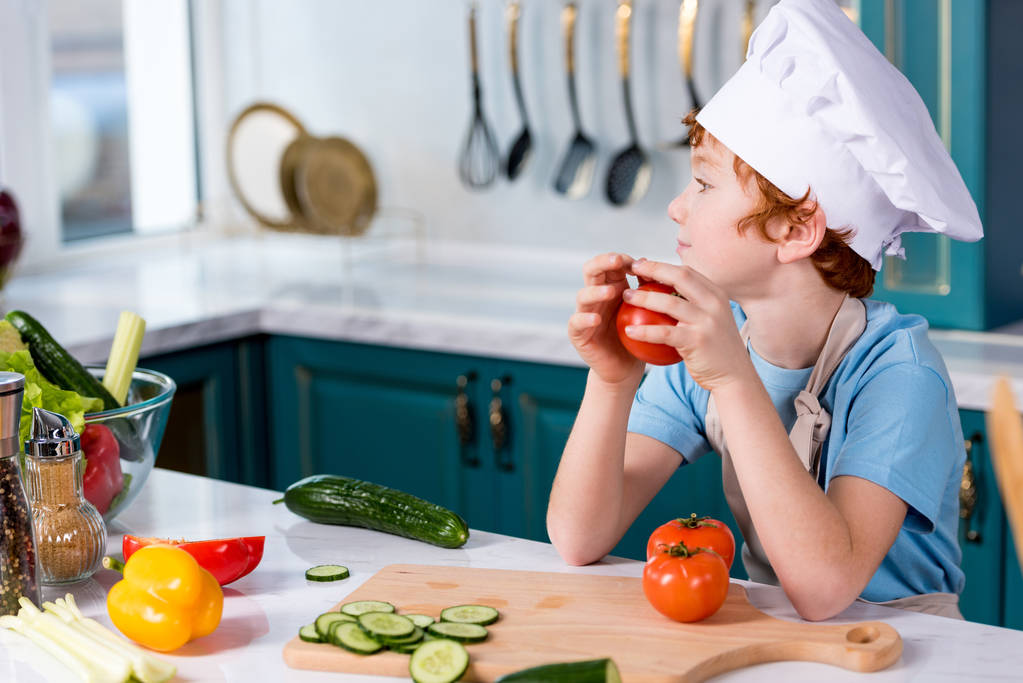 menino bonito em chapéu de chef e avental olhando para longe enquanto sentado na cozinha
 - Foto, Imagem