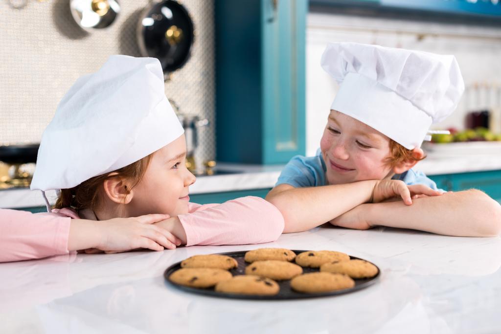 gelukkige kinderen in chef-kok hoeden glimlachen elkaar en heerlijke koekjes op tafel - Foto, afbeelding