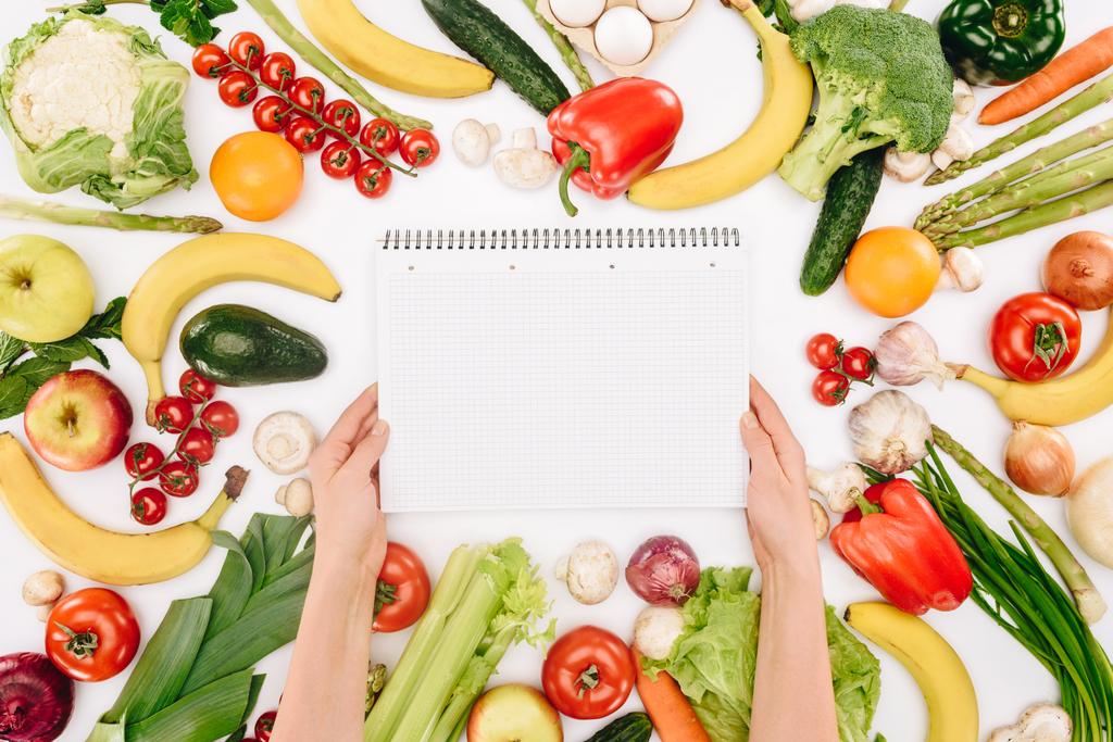 bijgesneden afbeelding van meisje holding laptop tussen groenten en fruit geïsoleerd op wit - Foto, afbeelding