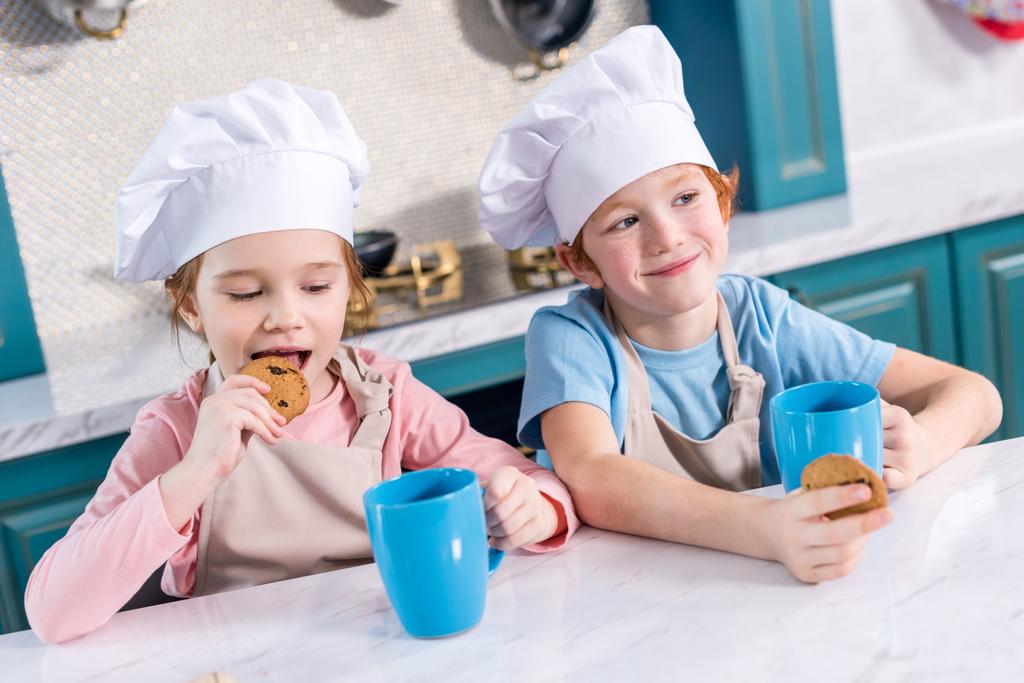 Παιδικά χαμόγελα σε καπέλα σεφ, πίνοντας τσάι και τρώει μπισκότα στην κουζίνα - Φωτογραφία, εικόνα