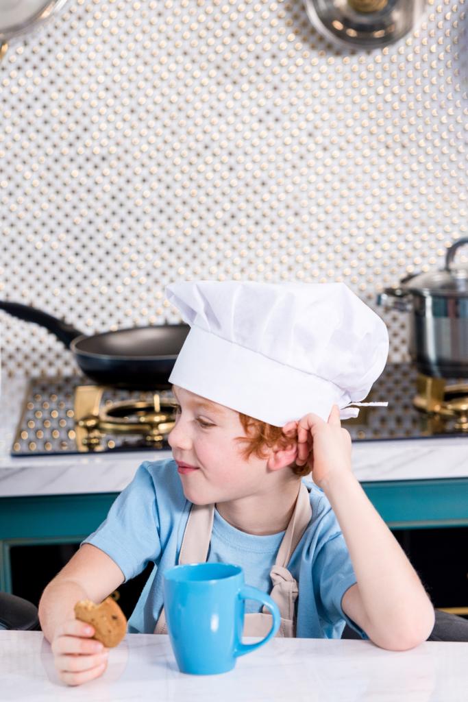 милый маленький мальчик в шляпе шеф-повара пьет чай и ест печенье на кухне
 - Фото, изображение