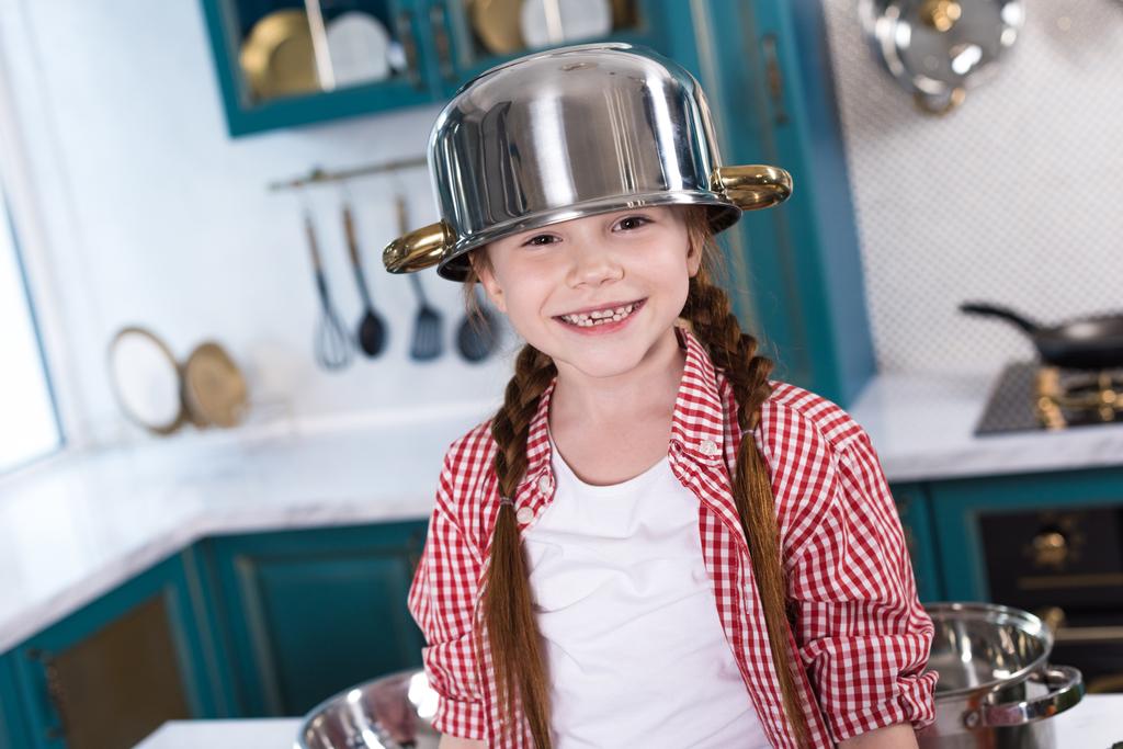 αξιολάτρευτο μικρό παιδί με την κατσαρόλα στο κεφάλι χαμογελώντας σε φωτογραφική μηχανή στην κουζίνα - Φωτογραφία, εικόνα