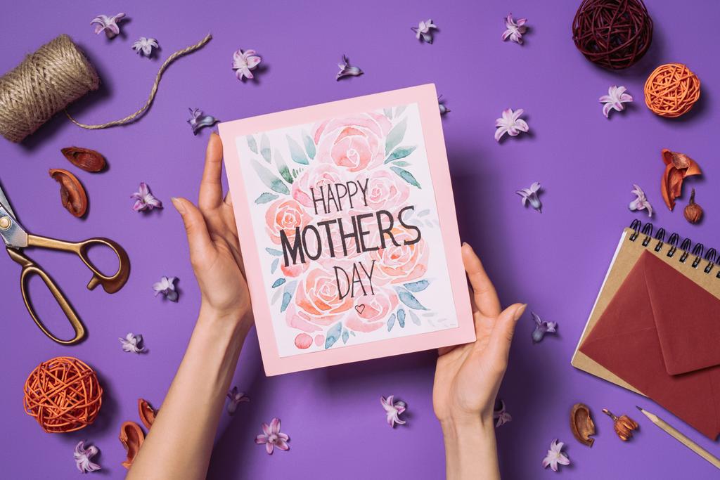 částečný pohled ženy držící šťastné matky den pohlednice v rukou s výzdobou kolem izolovaných na fialová - Fotografie, Obrázek