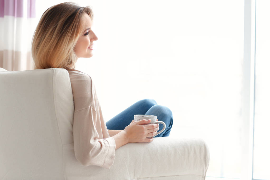 Šťastná mladá žena pít čaj, zatímco sedí v křesle u vás doma - Fotografie, Obrázek