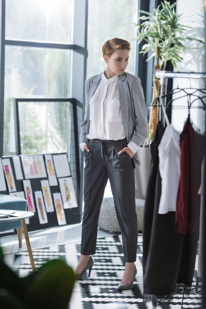 Junge Modedesignerin im modernen Büro mit vielen Kleidern auf Kleiderbügeln - Foto, Bild