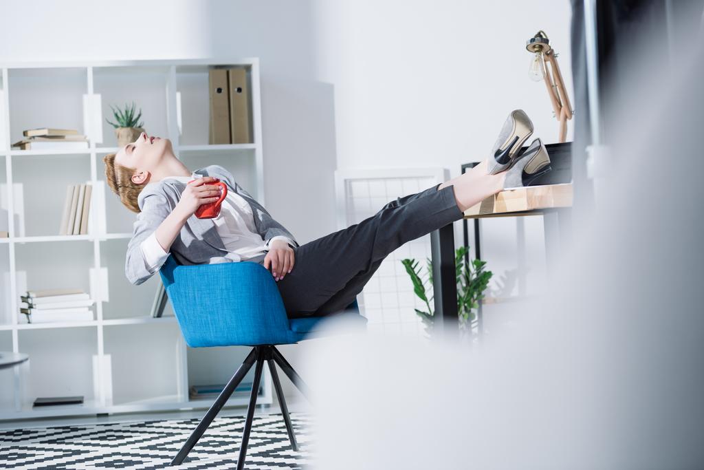 fashionable young businesswoman relaxing with cup of coffee in chair at office - Photo, Image