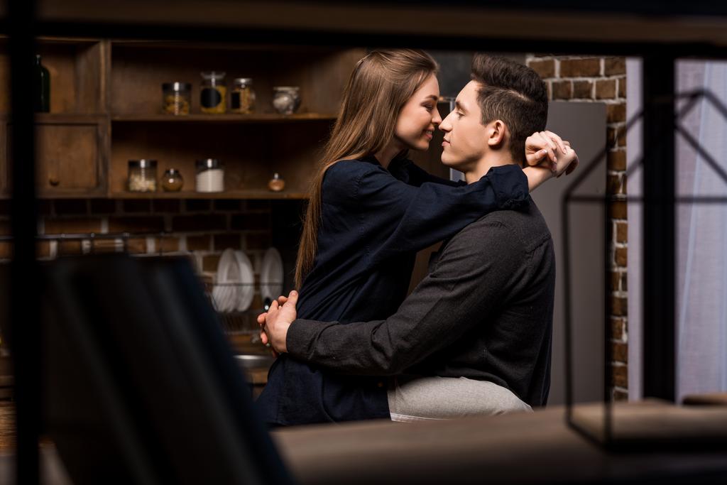 girlfriend sitting on table in kitchen and going to kiss boyfriend - Photo, Image