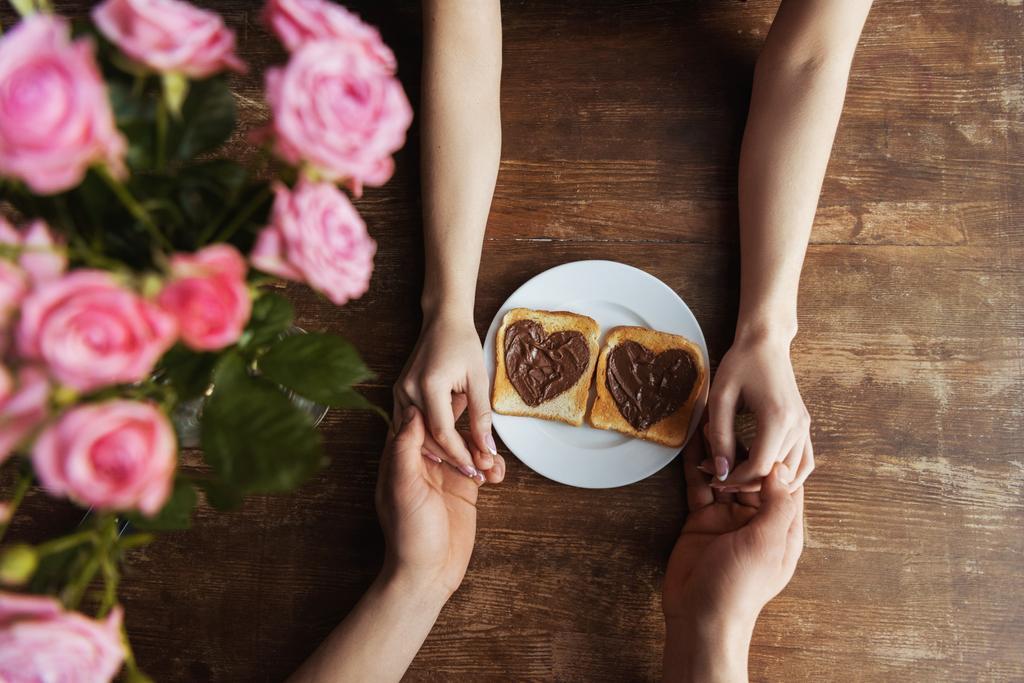 περικομμένη εικόνα ζευγάρι κρατώντας τα χέρια κοντά στο τοστ με πάστα σοκολάτας σε σχήμα καρδιάς, ημέρα του Αγίου Βαλεντίνου έννοια - Φωτογραφία, εικόνα