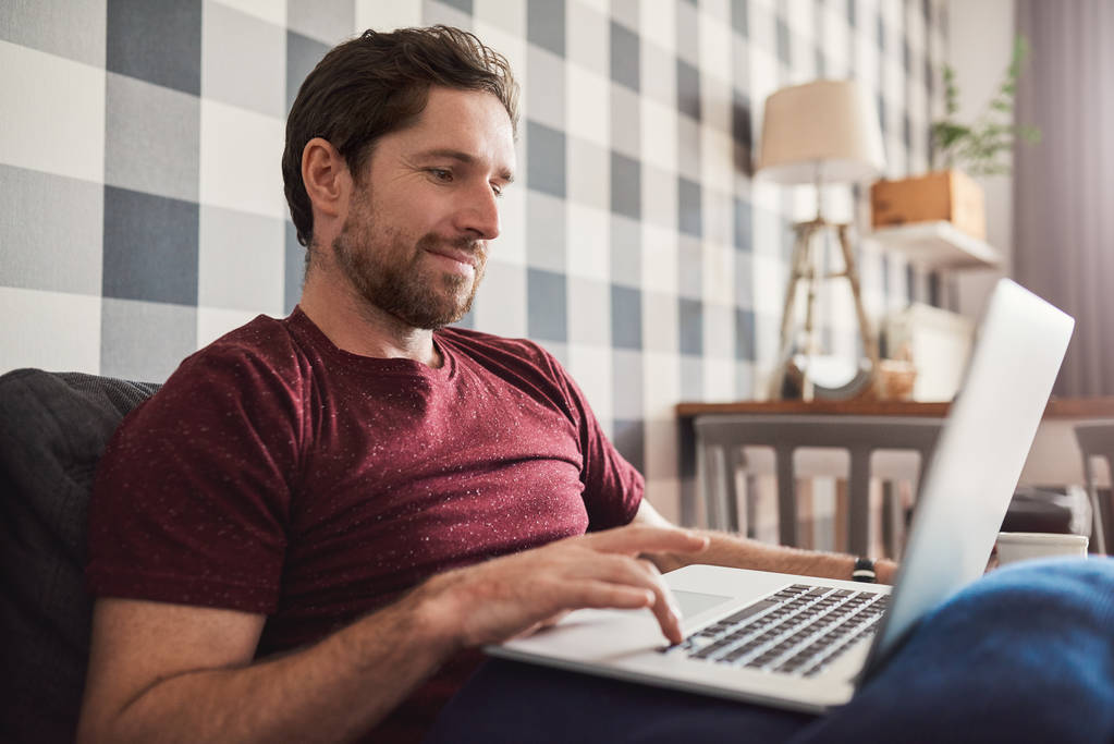 Sonriente joven navegando por Internet con el ordenador portátil mientras está sentado solo en su sofá de la sala de estar en casa
 - Foto, Imagen