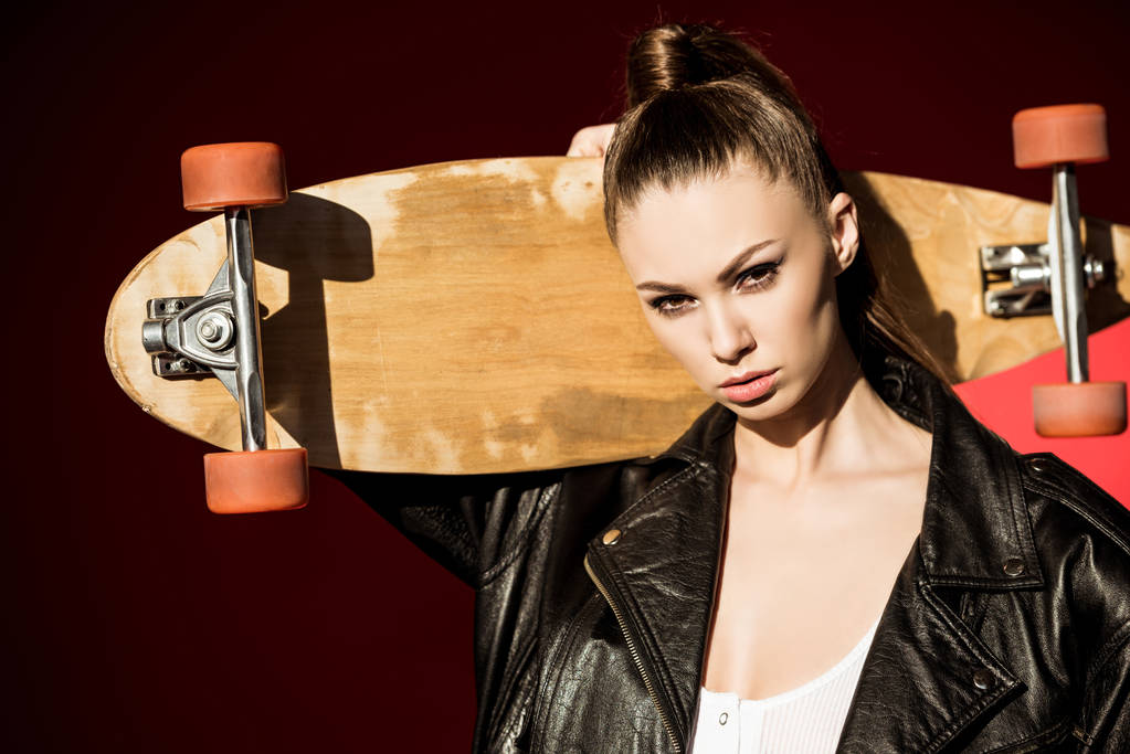 attractive fashionable girl in black leather jacket posing with longboard, on red - Photo, Image
