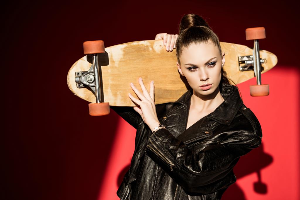 attractive girl in black leather jacket posing with longboard, on red - Photo, Image