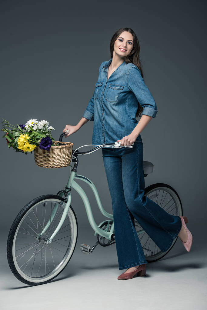 bella ragazza in abiti in denim che tiene in bicicletta con fiori in cesto di vimini, su grigio
 - Foto, immagini