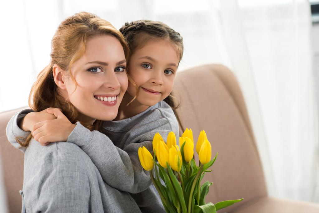 Gelukkig moeder en dochter met gele tulpen knuffelen en lachend op camera - Foto, afbeelding