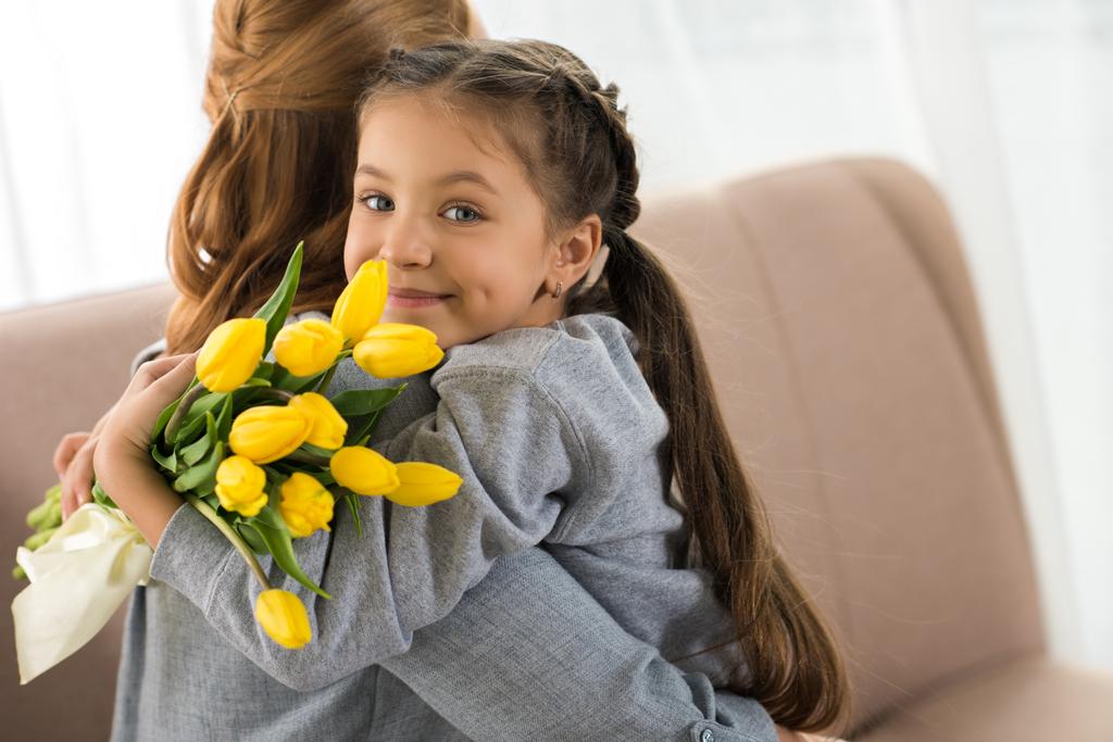 дитина з жовтими тюльпанами обіймає матір і посміхається на камеру
 - Фото, зображення