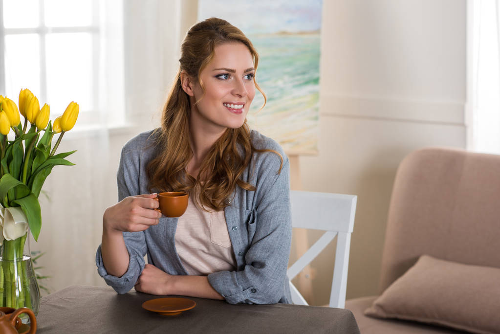 uśmiechający się młoda kobieta, trzymając kubek kawy i odwracając w domu - Zdjęcie, obraz
