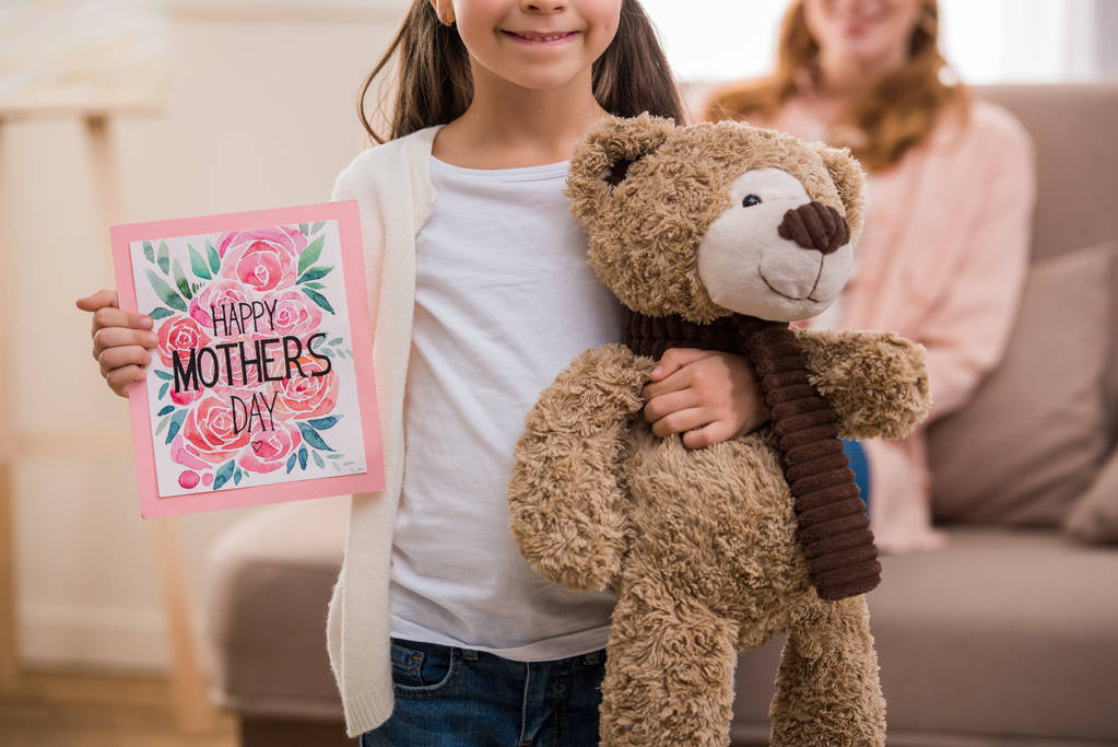 περικοπεί shot της ευτυχισμένη παιδί κρατώντας ευτυχισμένη μητέρες ημέρα ευχετήρια κάρτα και αρκουδάκι - Φωτογραφία, εικόνα