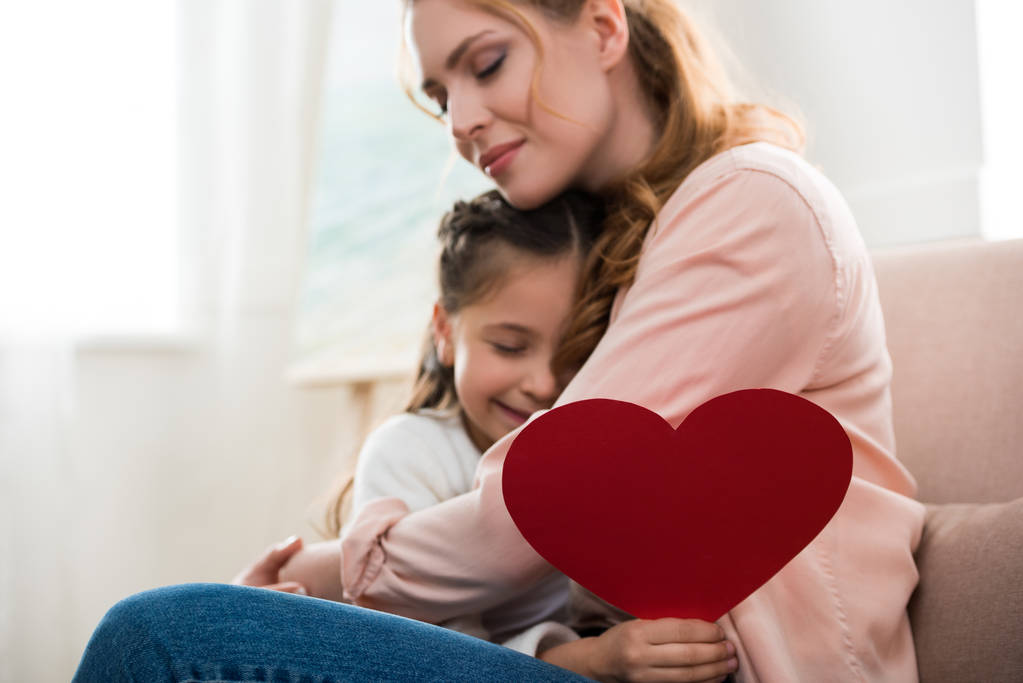 feliz madre e hija con el símbolo del corazón rojo abrazándose en casa
 - Foto, Imagen