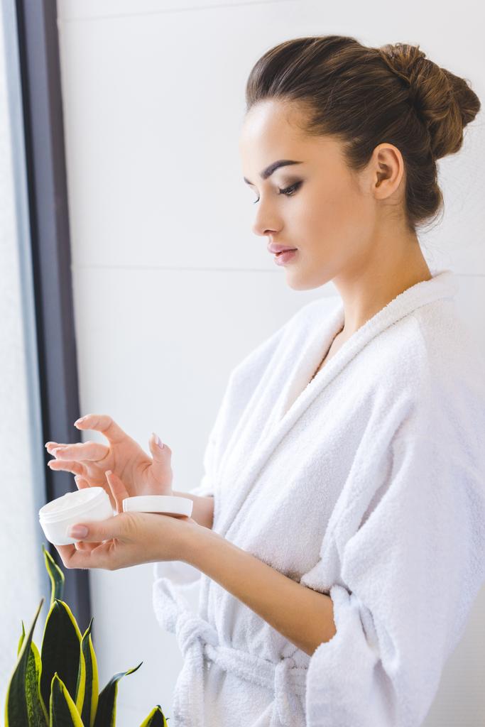 side view of beautiful woman in bathrobe with face cream in hands - Photo, Image