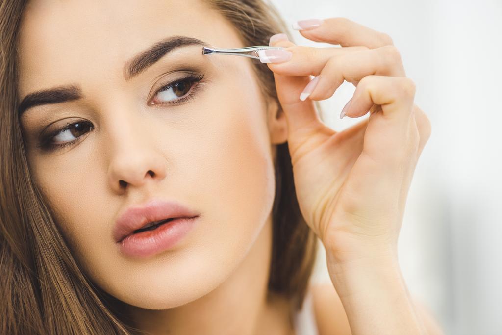 portrait of young woman plucking eyebrows with tweezers - Photo, Image