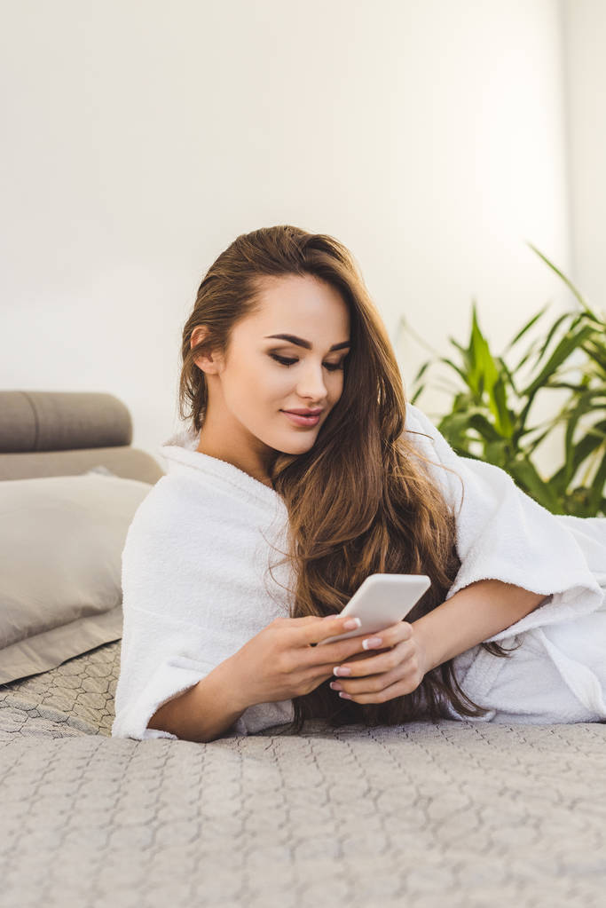 smiling young woman in bathrobe with smartphone lying on bed - Photo, Image