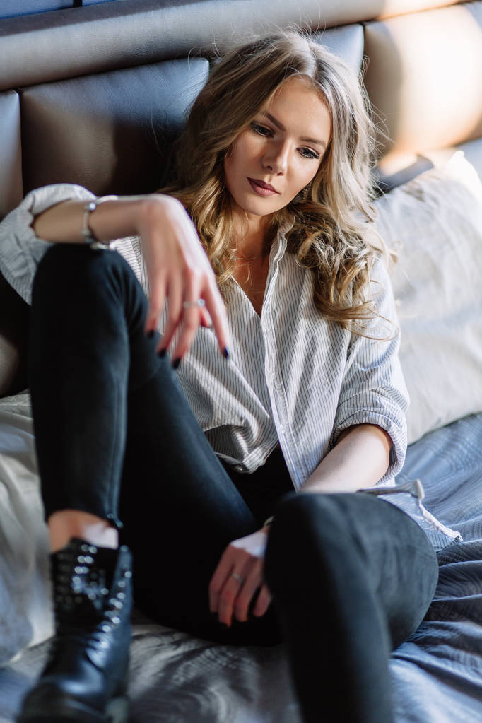 Een meisje met lang blond haar, gekleed in een gestreepte shirt, zwarte jeans en zwarte schoenen zitten op het bed met blauwe beddengoed. Modieuze casual outfit. - Foto, afbeelding