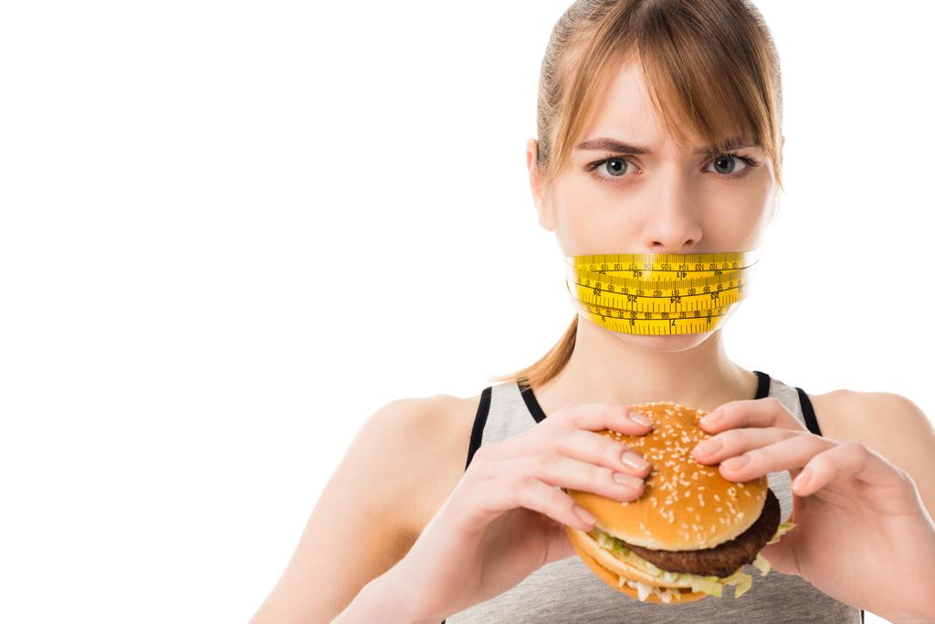 jeune femme avec la bouche attachée dans le ruban à mesurer tenant hamburger isolé sur blanc
 - Photo, image
