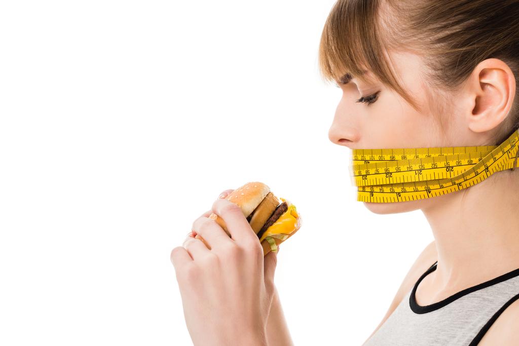 Frau mit im Maßband gefesseltem Mund versucht Burger isoliert auf Weiß zu essen - Foto, Bild