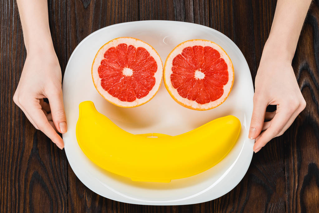 abgeschnittene Aufnahme einer Frau, die einen Teller mit halbierter Grapefruit und Bananenkoffer hält - Foto, Bild