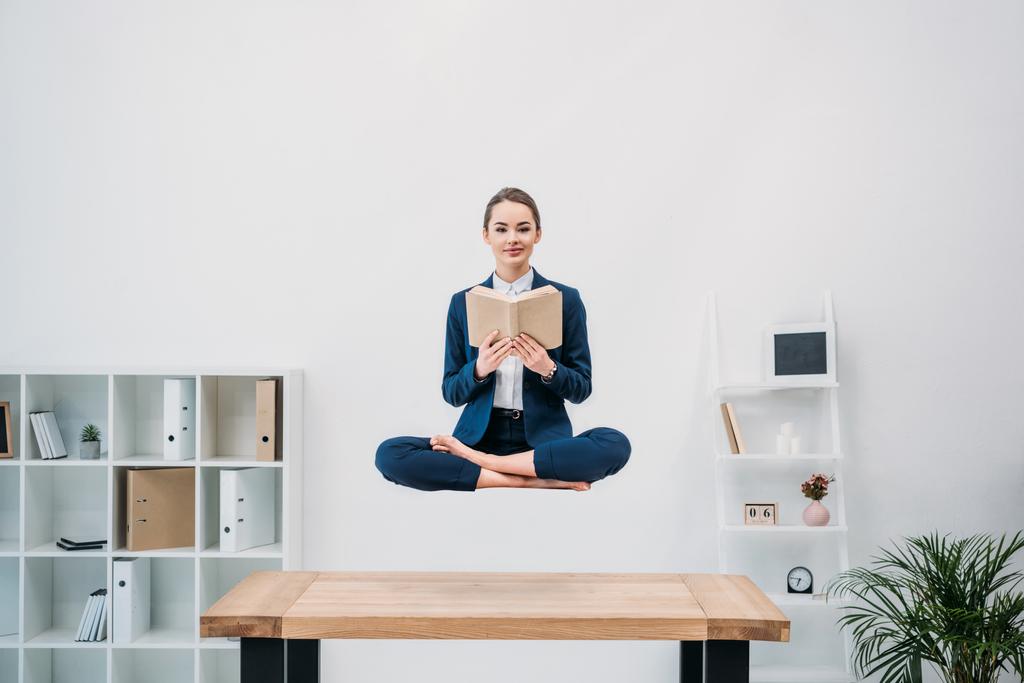smiling young businesswoman reading book while levitating at workplace  - Photo, Image