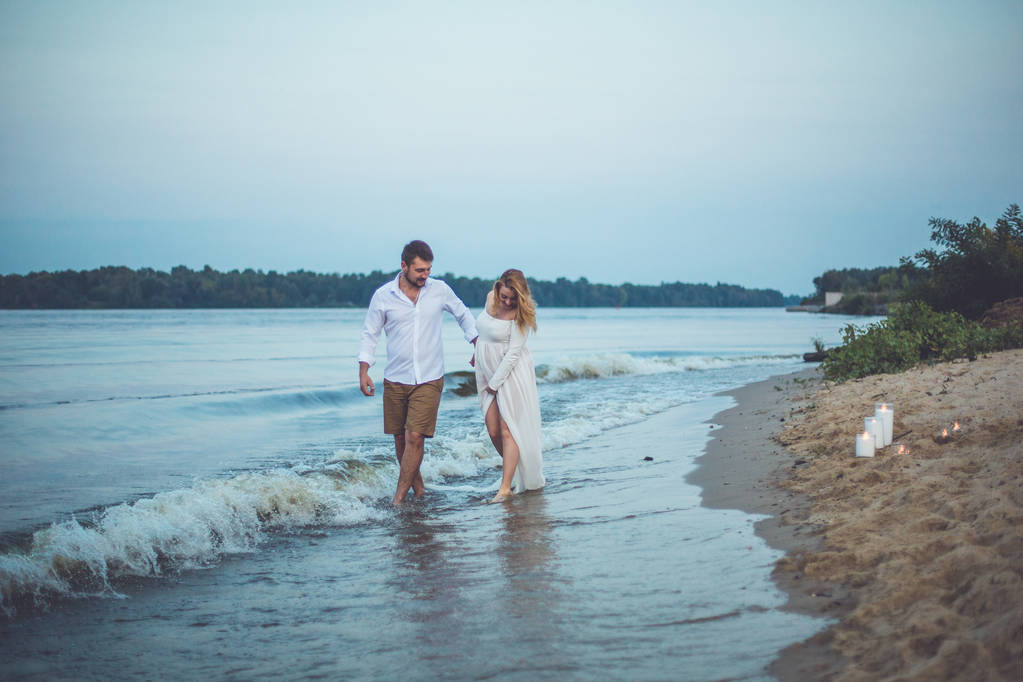 Giovane bell'uomo e sua moglie incinta che si tiene per mano e cammina sulla spiaggia del fiume
 - Foto, immagini