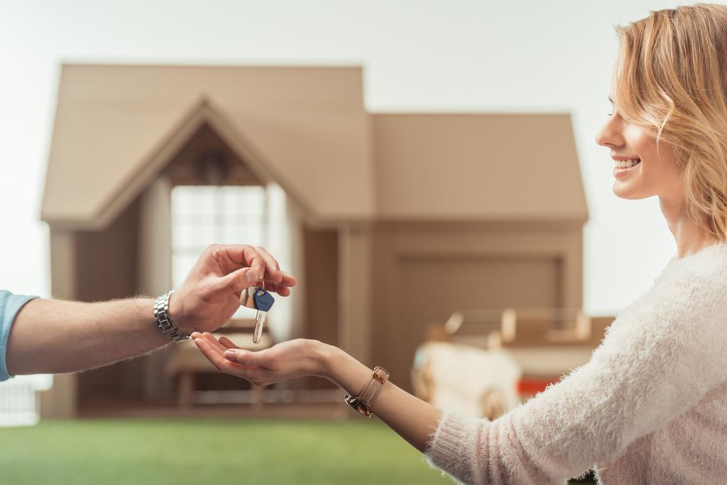обрезанный снимок агента по недвижимости передающий ключ счастливой женщине перед картонным домом
 - Фото, изображение