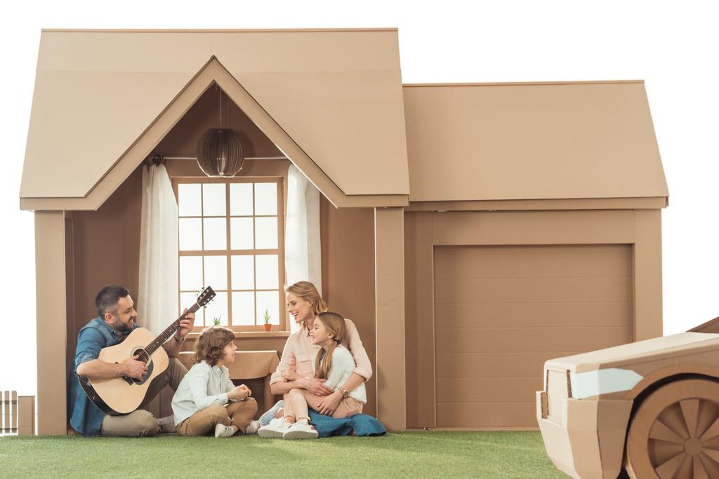 vader spelen gitaar voor kinderen en vrouw op nieuwe kartonnen huis geïsoleerd op wit - Foto, afbeelding