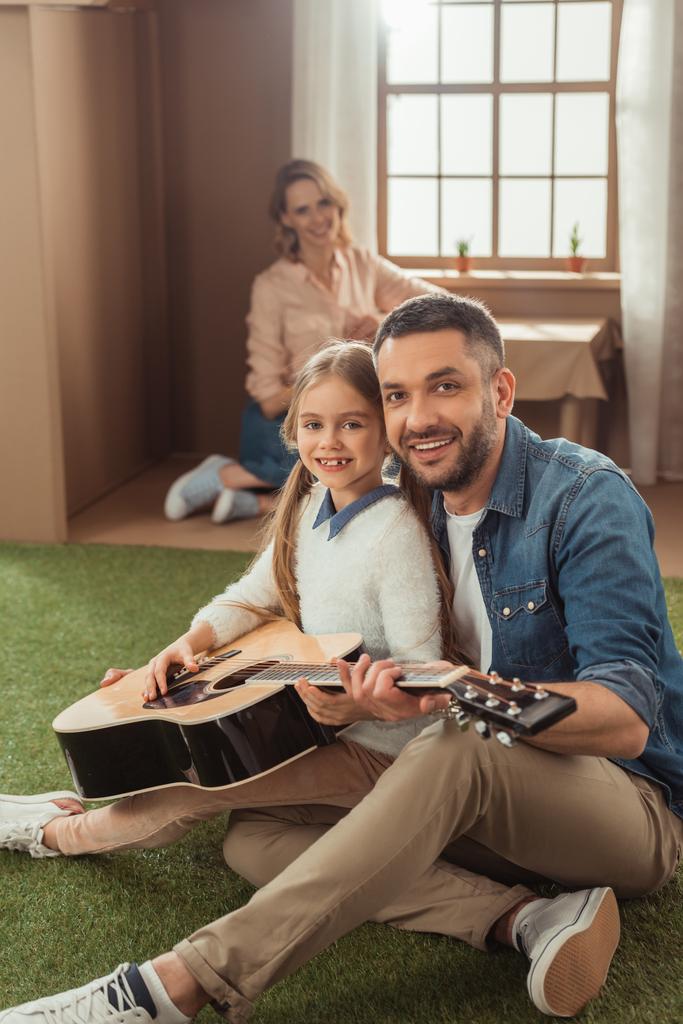 Gelukkig vader en dochter gitaar spelen op gras voor kartonnen huis met moeder zit in - Foto, afbeelding