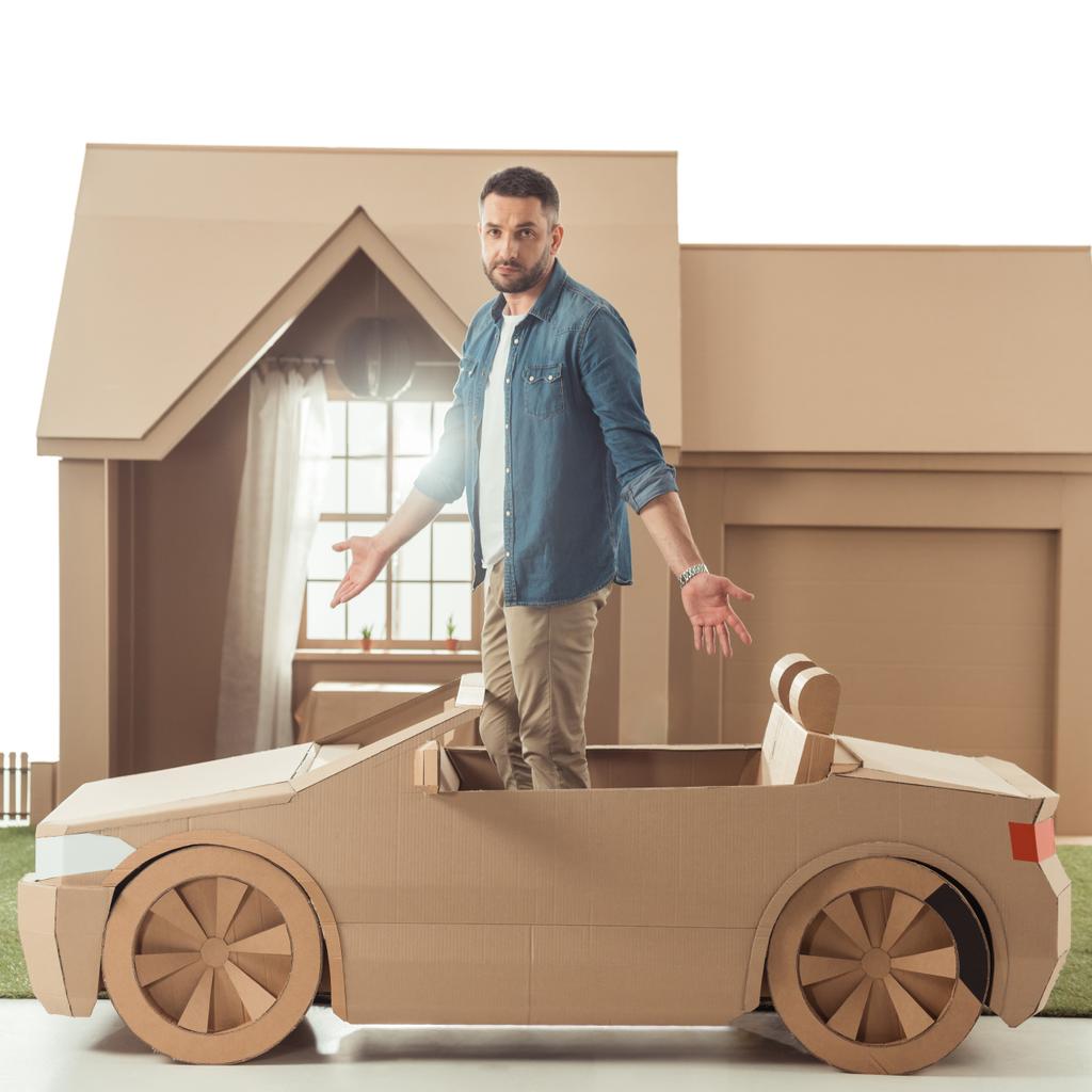 handsome man in cardboard car in front of cardboard house isolated on white - Photo, Image