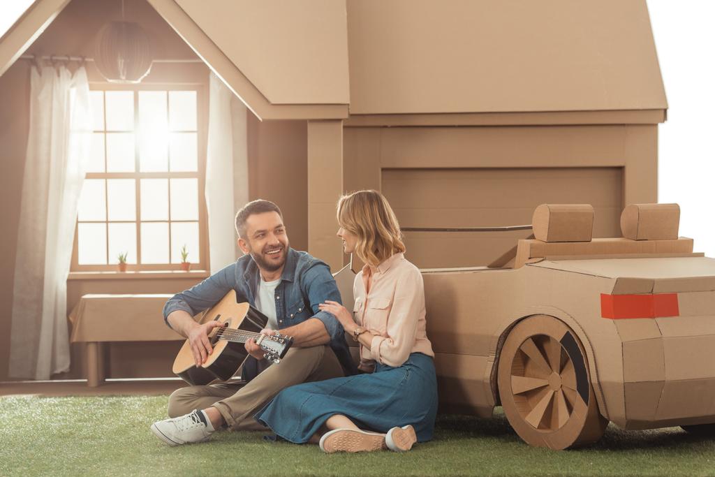man playing guitar for girlfriend on yard of cardboard house - Photo, Image