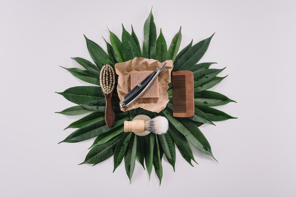 верхний вид организованных инструментов парикмахера для бритья на зеленых листьях, изолированных на сером
 - Фото, изображение