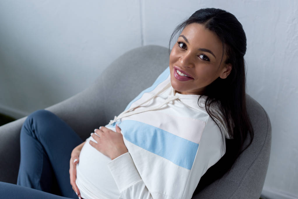 feliz mulher grávida afro-americana sentada em poltrona e olhando para a câmera
 - Foto, Imagem