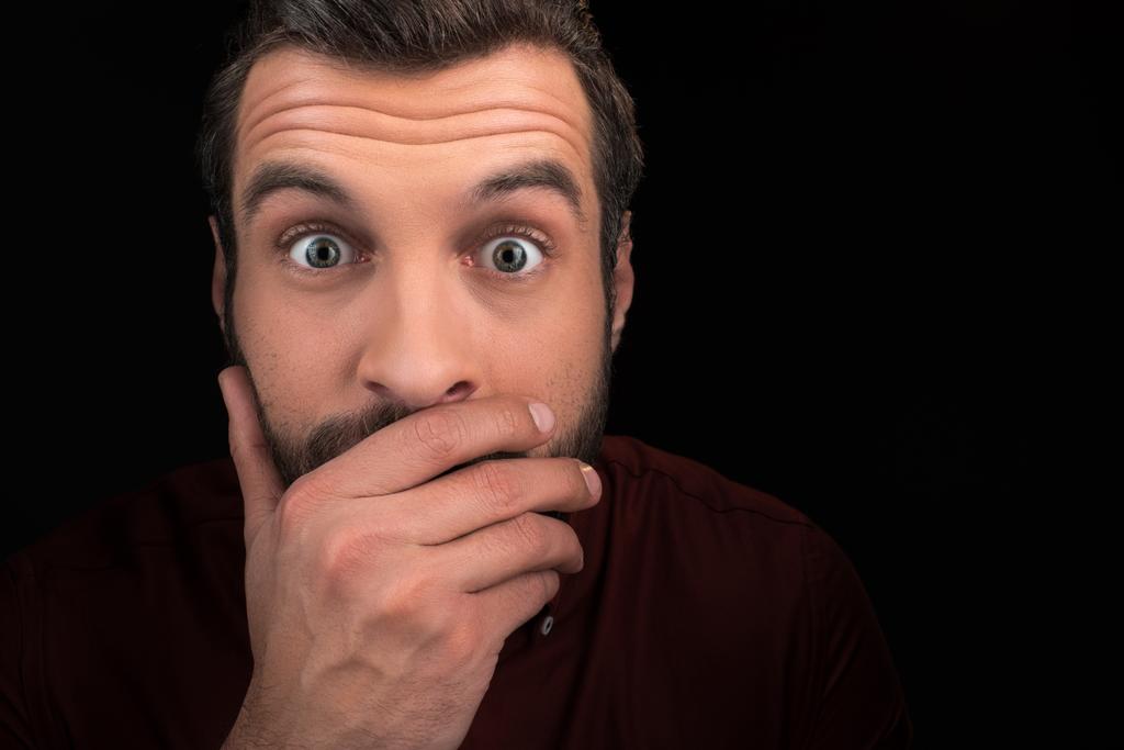 Porträt eines schockierten Mannes, der den Mund isoliert auf schwarz bedeckt - Foto, Bild