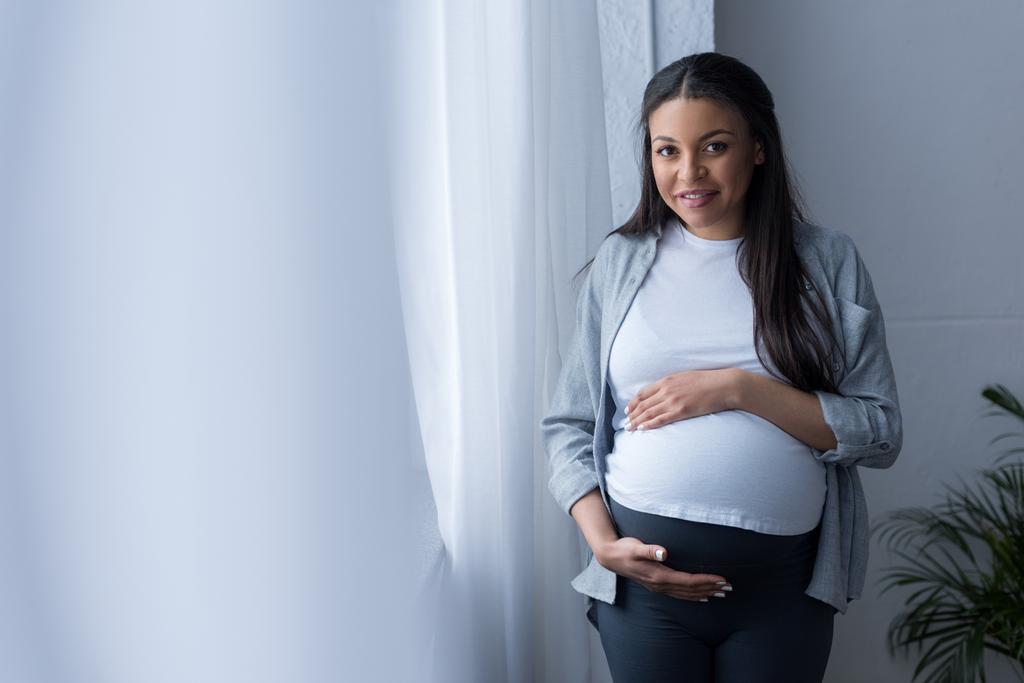 uśmiechający się african american Kobieta w ciąży dotykając jej brzuch, stojąc przy oknie - Zdjęcie, obraz