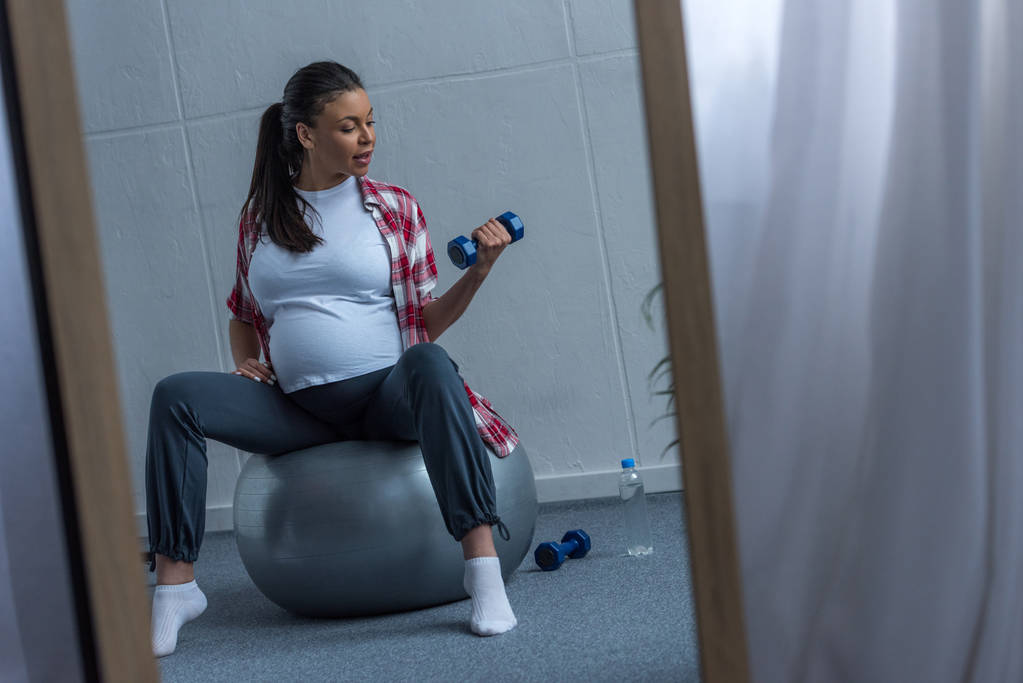 зеркало с отражением африканской беременной женщины, сидящей на мяче и тренирующейся с гантелями
 - Фото, изображение